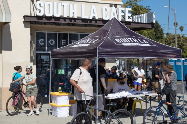 A crowd of bicyclists stopping by a pop-up tent for refreshments in front of a building with the sign "SOUTH LA CAFE".